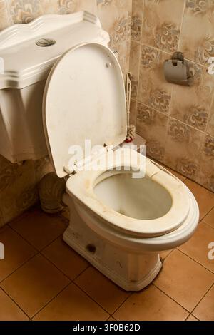dirty old and broken white toilet seat in tiled bathroom, stained ceramic bowl with open lid, angle view with copy space, poor hygiene and plumbing Stock Photo