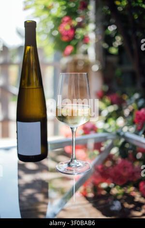White wine bottle next to a filled glass on a glass table, with vibrant flowers and greenery in a bright outdoor environment Stock Photo
