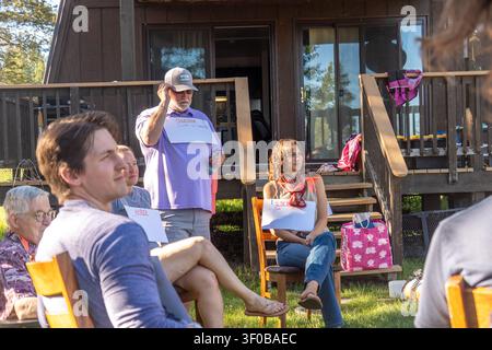 Man in light purple polo and cap delivers presentation from wooden cabin deck to seated audience. Name signs sit on chairs as attendees listen amid tr Stock Photo