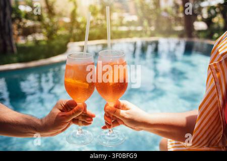 Two individuals clink glasses filled with orange cocktails by a swimming pool, with lush greenery in the background and sunlight reflecting on the wat Stock Photo