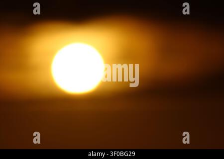 Telephoto close-up of the glowing sun partially hidden behind dark clouds during a warm golden sunset with atmospheric light. Stock Photo