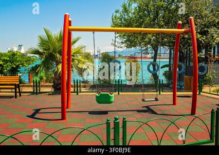 Empty colorful kids swing on safety rubber coating inside coastal public park facing turquoise sea under sunny sky. Stock Photo