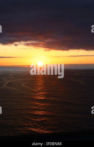 Bright orange sunset with the sun partially hidden behind clouds over the Atlantic Ocean, producing a vibrant evening seascape. Stock Photo