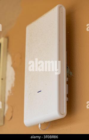 A close-up view of a white outdoor wireless access point device attached to a textured orange wall, showing signs of dust accumulation and a connected Stock Photo