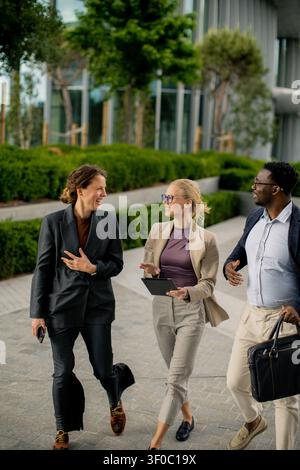 Three professionals are walking together along a path next to a modern building. They are engaged in conversation and appear to be enjoying their disc Stock Photo