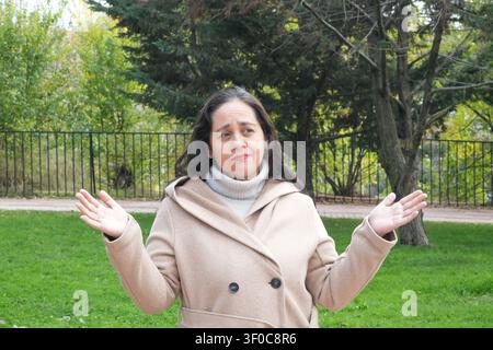 Woman gestures with open hands outdoors. She wears a beige coat and turtleneck. Green grass and trees form the background. Fence lines the park behind Stock Photo