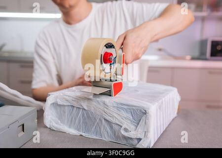 Person using tape dispenser to securely wrap cardboard box in plastic wrap on home desk, preparing parcels for shipping, moving, small business orders Stock Photo
