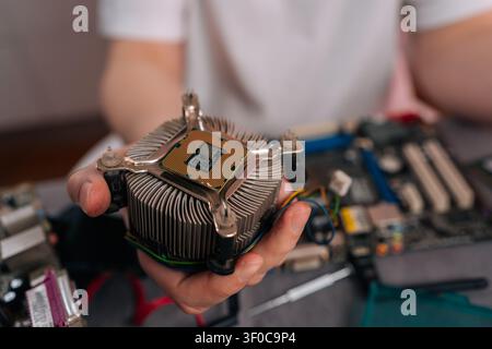 Hand holding CPU mounted on heatsink above computer motherboard, ready for installation or repair close-up of technician performing hardware Stock Photo