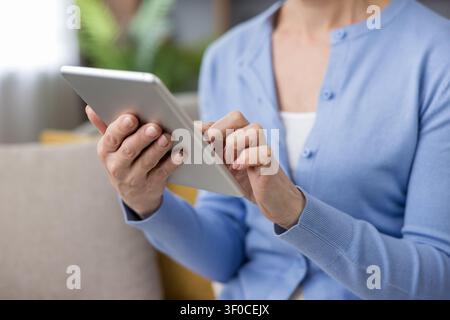 Hands of an older woman scrolling and tapping on a modern digital tablet, connecting with friends and family or enjoying online content at home Stock Photo