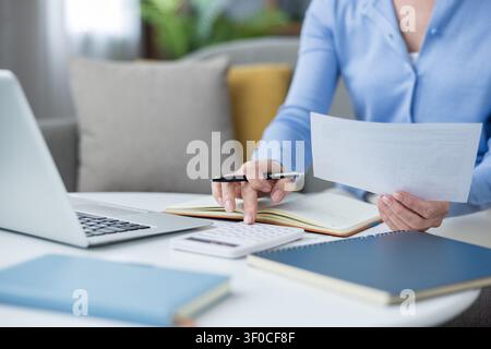 Woman's hands using a calculator, holding a document, and noting details in a notebook on a white table with a laptop, managing personal finances from her home office Stock Photo