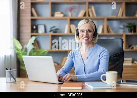 Smiling middle aged woman wearing a headset and spectacles using a laptop while working productively from her home office, representing online communication and customer service Stock Photo