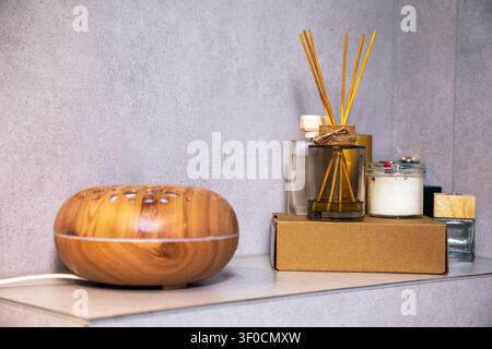 Cozy bathroom decor with electric wooden oil diffuser, fragrance reed sticks, perfume bottles, and burning candles on a shelf for relaxation Stock Photo