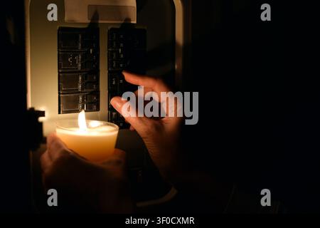 Close-up side view showing hand flipping circuit breaker switch in electrical panel while holding lit candle for illumination during blackout Stock Photo