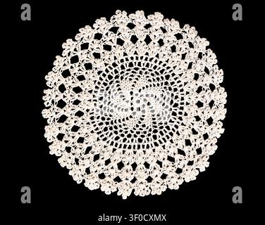 Delicate white crocheted doily featuring intricate floral lace pattern and scalloped edge isolated cutout on black background Stock Photo