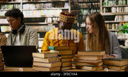 Students happily discussing study materials and reading from textbooks at shared library table, collaborative work during group study surrounded by green lamps and bookshelves. Stock Photo