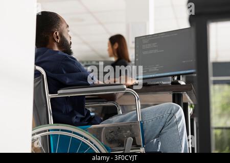 Male wheelchair user works at desktop PC, debugging software and reviewing website code. Dedicated black man developer completes programming tasks in inclusive tech startup environment. Stock Photo