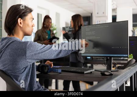 Asian male programmer points at desktop monitor with surprised expression after identifying key code statement to solve web development issue. Man reviews HTML scripts in modern tech workspace. Stock Photo