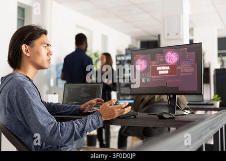 Young programmer having a stressed expression while seeing a critical error message and data breach pop ups on his computer monitor, indicating a serious cyber attack or system failure Stock Photo