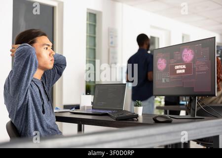 Young adult male developer feeling frustrated and stressed, watching an alarming critical error message displayed on his monitor while working on coding in modern technical workspace. Stock Photo
