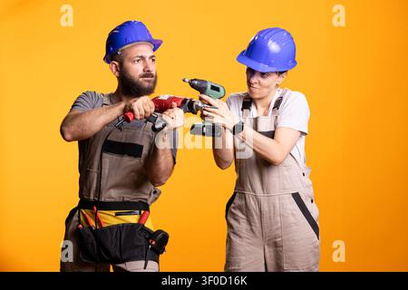 Male and female builders take break from work, sharing playful moment with battery operated drills. Construction workers wearing safety helmets, playfully pose with power tools in modern studio. Stock Photo