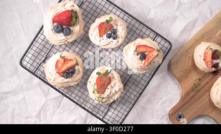 Five individual mini pavlovas topped with fresh strawberry slices, blueberries, and herb sprigs are arranged on a black wire rack, showcasing a crisp, Stock Photo