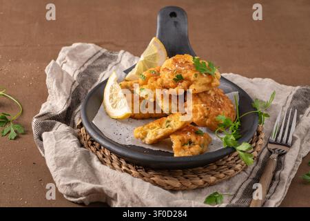 Crispy, golden-brown cod fish fritters are arranged on parchment paper in a dark skillet, garnished with fresh lemon wedges and parsley. Soft overhead Stock Photo