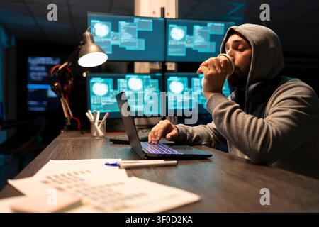 Caucasian man in hoodie drinks caffeine, monitoring encrypted data streams on device screen. Male developer works late at desk, checking code and debugging on laptop with coffee in office. Stock Photo
