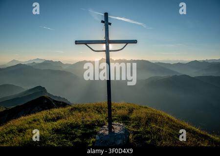 The rays of the setting sun filter from behind the cross creating an effect of divine light and spirituality that envelops the surrounding peaks Stock Photo