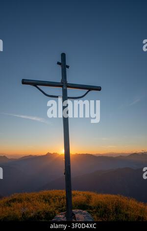 The rays of the setting sun filter from behind the cross creating an effect of divine light and spirituality that envelops the surrounding peaks Stock Photo