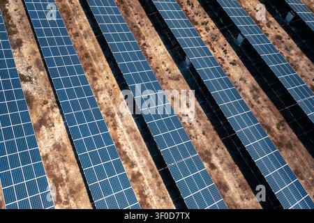 High angle aerial photo showing multiple rows of ground mounted solar panels on dry brown terrain, with bright sunlight reflections, repeating grid structure, shadow gaps, and expansive clean energy Stock Photo
