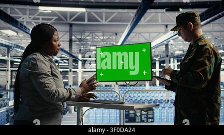 Soldier and female technician use a green screen computer in a server hub to manage systems with a mockup workstation. Stock Photo