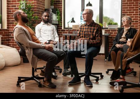 Diverse people sit in a circle at an addiction support meeting, talking through personal issues during a mental health and rehabilitation therapy session. Stock Photo