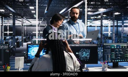 Team of engineers brainstorming in server room working with AI to check digital infrastructure diagnostics, while teamworking IT team members in server facility assessing data using AI tools. Stock Photo