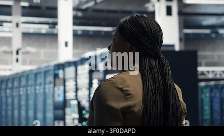 Woman walking through server facility rows of servers, checking equipment for system stability. IT professional supervising server environment, checking infrastructure mainframes. Stock Photo