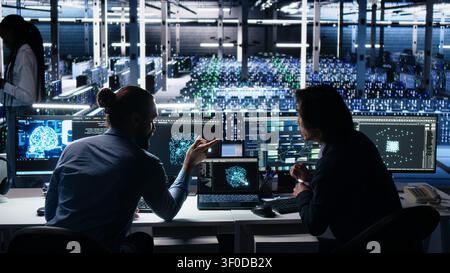 Senior programmer and coworker programs on a computer workstation in a data center with generative AI assistance. Stock Photo