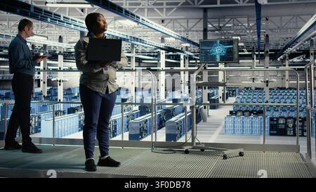 Server facility supervisor on industrial platform working to improve AI systems processing speed. African American server facility supervisor supervising equipment supporting AI workloads. Stock Photo