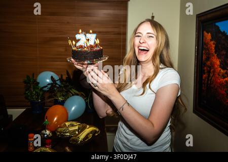 Smiling woman presenting chocolate birthday cake with lit candles at indoor party Stock Photo