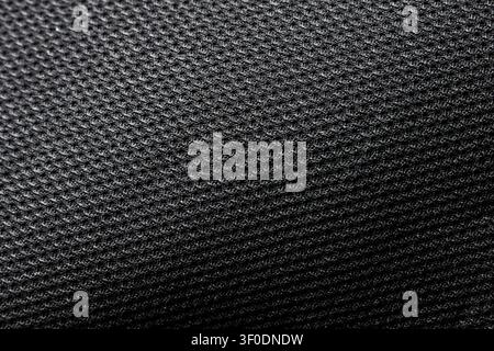 A black textured fabric surface with repeating small honeycomb knit pattern and soft directional light creating subtle highlights and shadow, close up Stock Photo