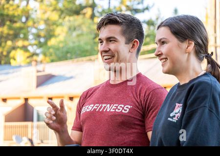 Two friends share candid moment outdoors, smiling and talking as warm sunlight washes over casual fitness shirts, with relaxed body language, soft bac Stock Photo