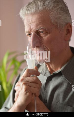 elderly man calmly using inhaler in indoor setting with plant in background side view. healthcare and elderly care context. gray and green tones Stock Photo
