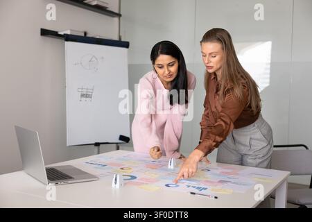 Businesswomen discussing marketing data, pointing at graphs leaning over table Stock Photo