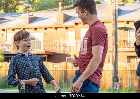Two men share light conversation in sunny backyard setting beside wooden house, with smiling faces, casual clothing, green grass, fence, and relaxed n Stock Photo