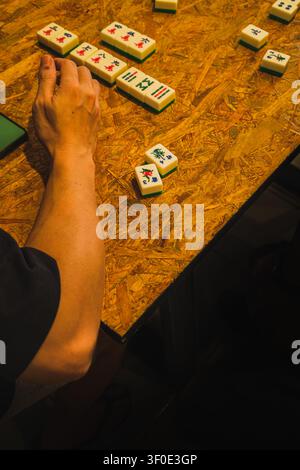 A high-angle close-up view of a player's arm and hand on a wooden particle board table, next to sections of exposed and discarded Mahjong tiles during Stock Photo