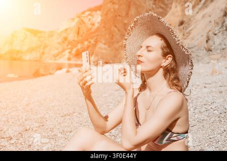 Woman, beach, makeup, applying lipstick while looking at her smartphone screen on a rocky shore during a warm summer day Stock Photo