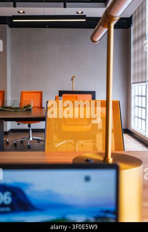 Modern office interior with orange mesh chairs, wooden desks, windows, and a laptop in the foreground. Stock Photo