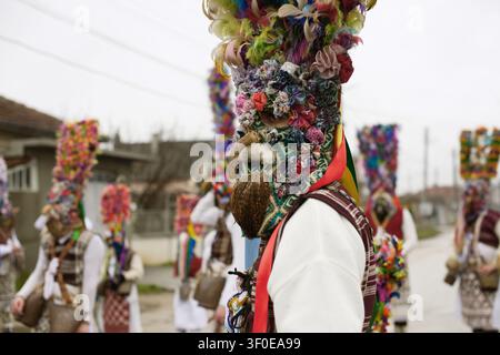 March 2, 2025 - Kalipetrovo, Bulgaria: A participant in the local Grebentsi kukeri tradition wears a tall decorated mask and headpiece with multicolor Stock Photo