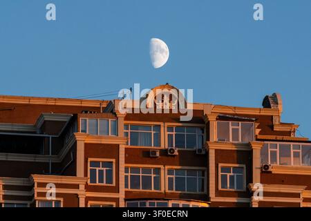 View of a residential building with a crown-shaped architectural detail on the roof under a clear blue sky with a visible half moon in Aktau, Mangysta Stock Photo