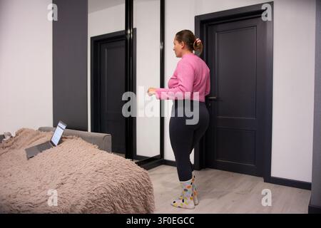 In a bedroom, a woman in a pink sweater and leggings stands near a bed with a laptop. The modern interior is highlighted by sleek, black doors and neu Stock Photo