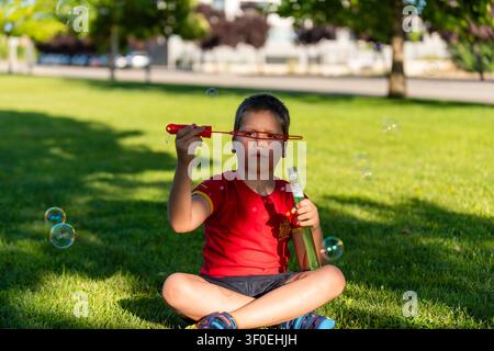 A boy sits on the grass in a park, happily blowing bubbles with a bubble wand during a sunny afternoon. Stock Photo