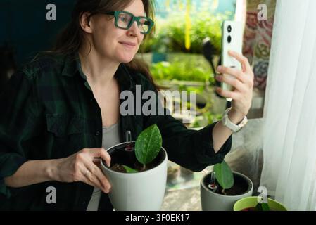 A proud woman takes a selfie with a potted plant in a sunny cozy home. The happy plant parent smiles holding indoor greenery and seedling in a flowerpot showing joyful excitement by the window. Stock Photo
